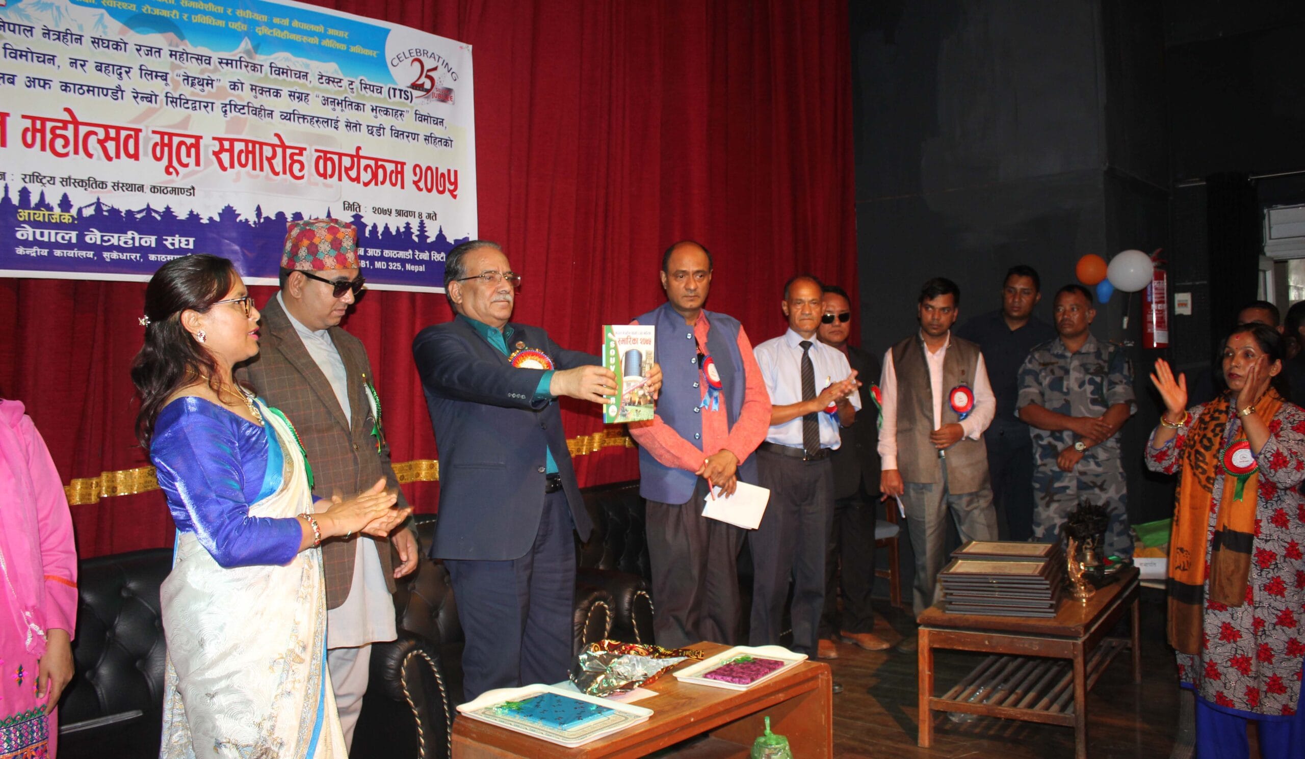 A formal event with a banner in the background celebrating the Silver Jubilee of the Nepal Association of the Blind (NAB). Several dignitaries are on stage, including a man holding up a book, while others clap and look on. A woman in a saree is seen addressing the gathering.