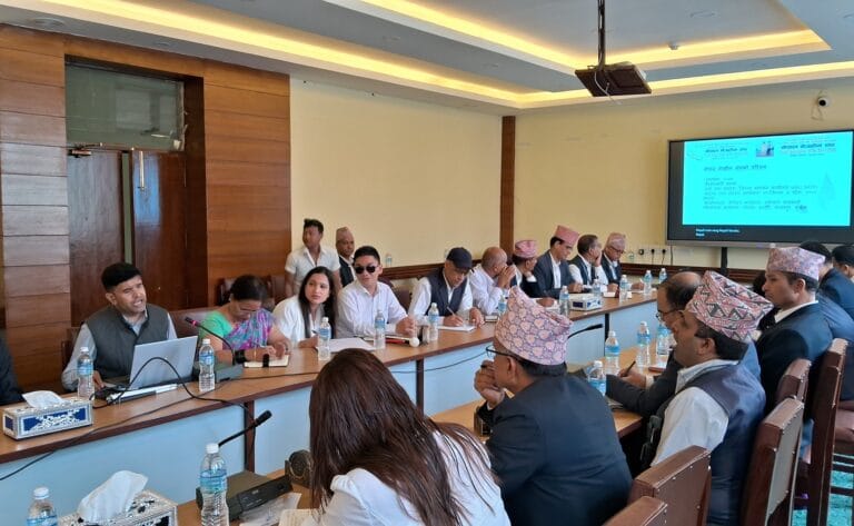A meeting room with participants seated around a long table. Several individuals, including men in traditional Nepali caps, are actively participating in a discussion. A presentation slide is visible on a screen in the background.