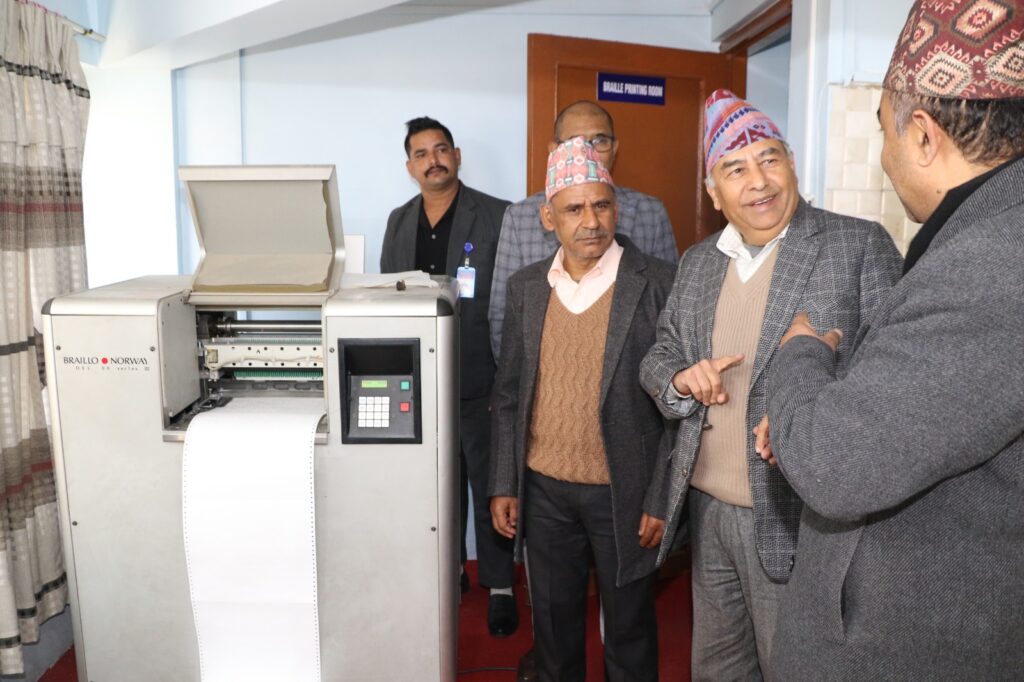 प्रमुख अतिथिज्यू र अन्य सहभागीहरू ब्रेल प्रिन्टिङ मेसिनको अवलोकन गर्दै गरेको दृश्य। सहभागीहरू मेसिनको कार्यप्रणाली र प्रयोगका बारेमा जानकारी लिँदै छन्।