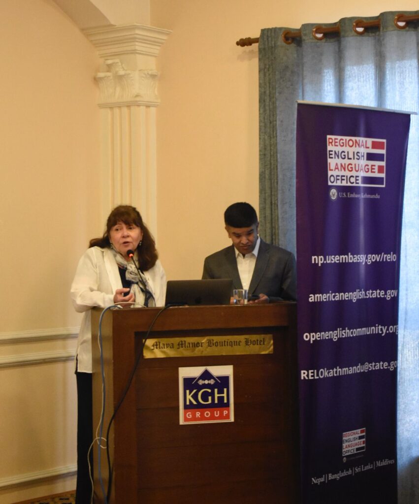 A medium shot of two presenters standing at a wooden podium during the training program. On the left, a woman in a white blazer and patterned scarf addresses the audience through a podium-mounted microphone. Beside her, a man in a grey suit jacket looks down toward a laptop on the lectern. The podium features the "Maya Manor Boutique Hotel" and "KGH Group" logos. To the right, a tall purple banner for the Regional English Language Office (RELO) is prominently displayed, listing official websites and contact information for RELO Kathmandu.