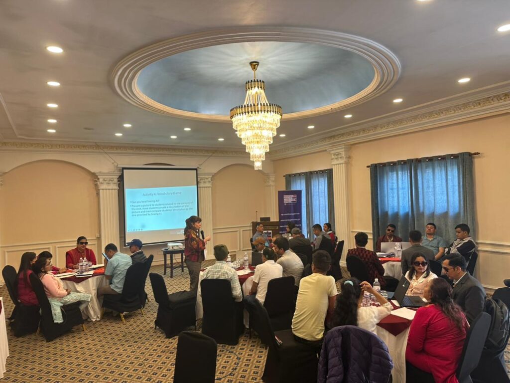 An overhead, wide-angle view of a professional training workshop taking place in a well-lit hotel conference room. Approximately twenty participants are seated at several round tables, engaged in group discussions and activities. A female facilitator stands at the front of the room near a large projection screen displaying a presentation titled "Activity 4: Vocabulary Game." The room features elegant decor, including a large tiered crystal chandelier hanging from a recessed circular ceiling. To the side, a blue RELO brand banner is visible. The atmosphere is one of active learning and collaboration among the English language teachers.