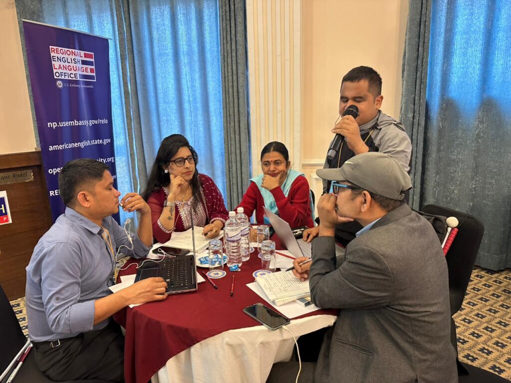 A group of five participants are gathered around a circular table covered with a maroon tablecloth at an event hosted by the Regional English Language Office (RELO). On the left, a man in a light blue shirt works on a laptop. To his right, two women and another man in a grey jacket and cap listen intently, some taking notes. Standing behind them, a male participant speaks into a handheld microphone, addressing the group or the room. The background features a tall blue banner with the RELO logo and U.S. Embassy web addresses, alongside light-colored curtains and patterned carpeting.