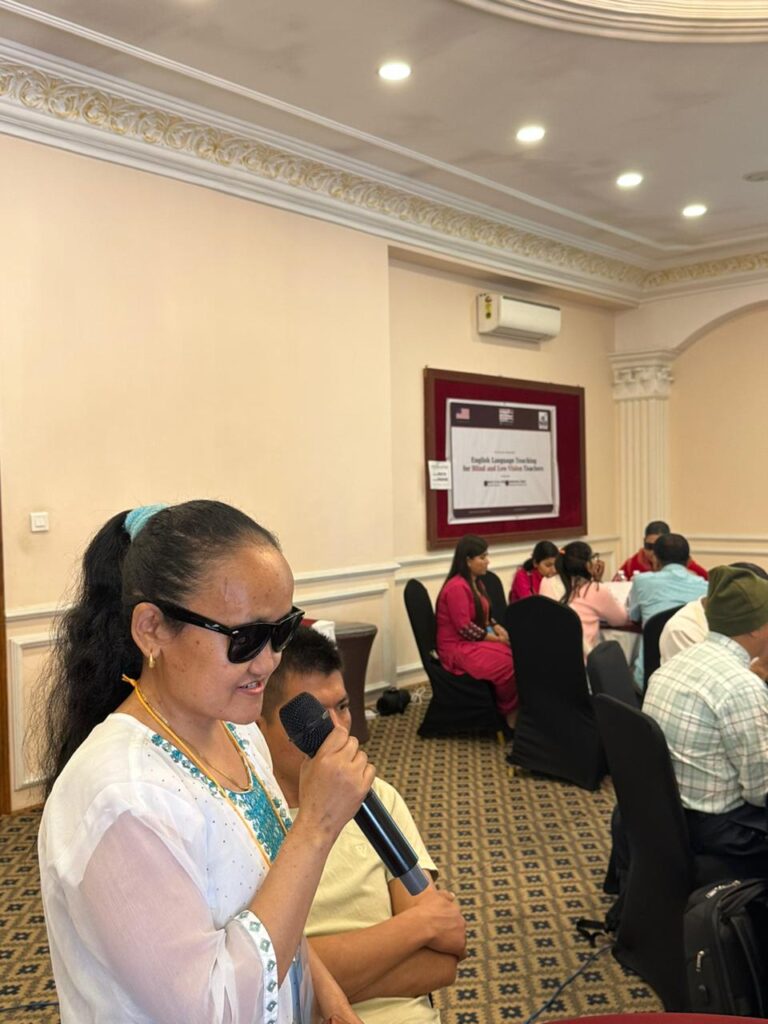A medium shot of a female participant with dark sunglasses speaking into a handheld microphone during a workshop. She is wearing a white top with blue embroidered details and has her hair tied back. In the background, other participants are seated at round tables, and the event banner for the "English Language Teaching for Blind and Low Vision Teachers" program is visible on the wall. The setting is a brightly lit conference room with ornate ceiling molding and patterned carpet, capturing a moment of inclusive discussion and experience sharing.