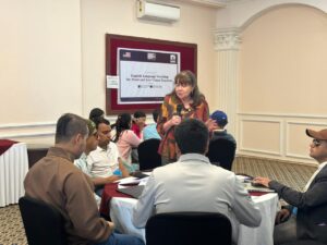 Prof. Madaline is speaking with a microphone during a training session while participants sit around tables and listen. Behind her, a banner reads “English Language Teaching for Blind and Low Vision Teachers,” indicating a training program held in Kathmandu, Nepal. Several participants, including individuals wearing name tags, are seated in groups with papers and materials on the table, suggesting a workshop or interactive learning activity.