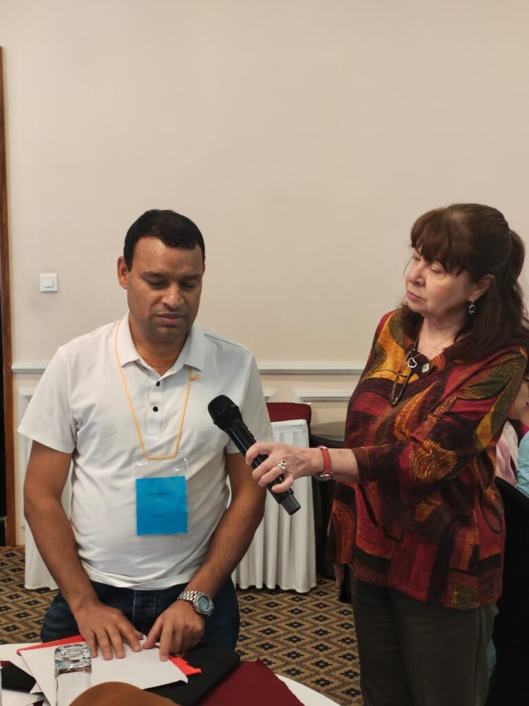 A medium shot showcasing an interactive moment during the training workshop. On the right, a female facilitator in a patterned red and orange blouse holds a microphone for a male participant seated on the left. The participant, wearing a white polo shirt and a blue identification badge, has his hands placed on a braille document or tactile teaching material on the table. The facilitator watches attentively as he prepares to speak. The background is a clean, neutral-toned wall, focusing the viewer's attention on the exchange and the inclusive nature of the educational setting.