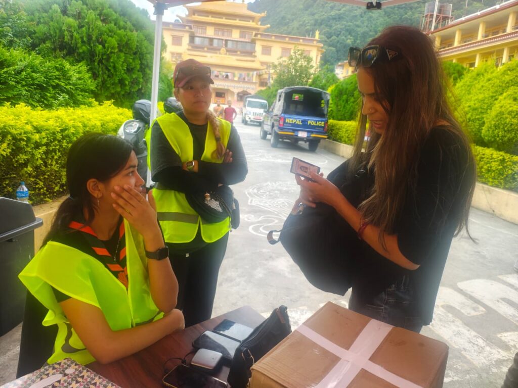 An outdoor registration or check-point setting where Ms. Paru Sharma (Country Coordinator of NAB) stands holding an identification card while interacting with two staff members of Yellow Gumba wearing high-visibility vests seated at a desk.