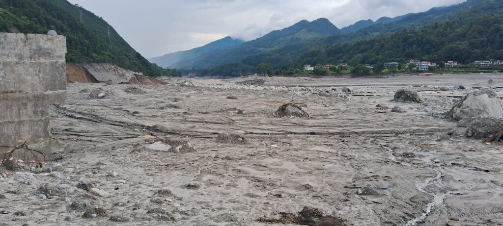 An outdoor landscape showing a wide, sediment-covered riverbed flanked by hills on the left and a damaged multistory building structure along the riverbank on the right.