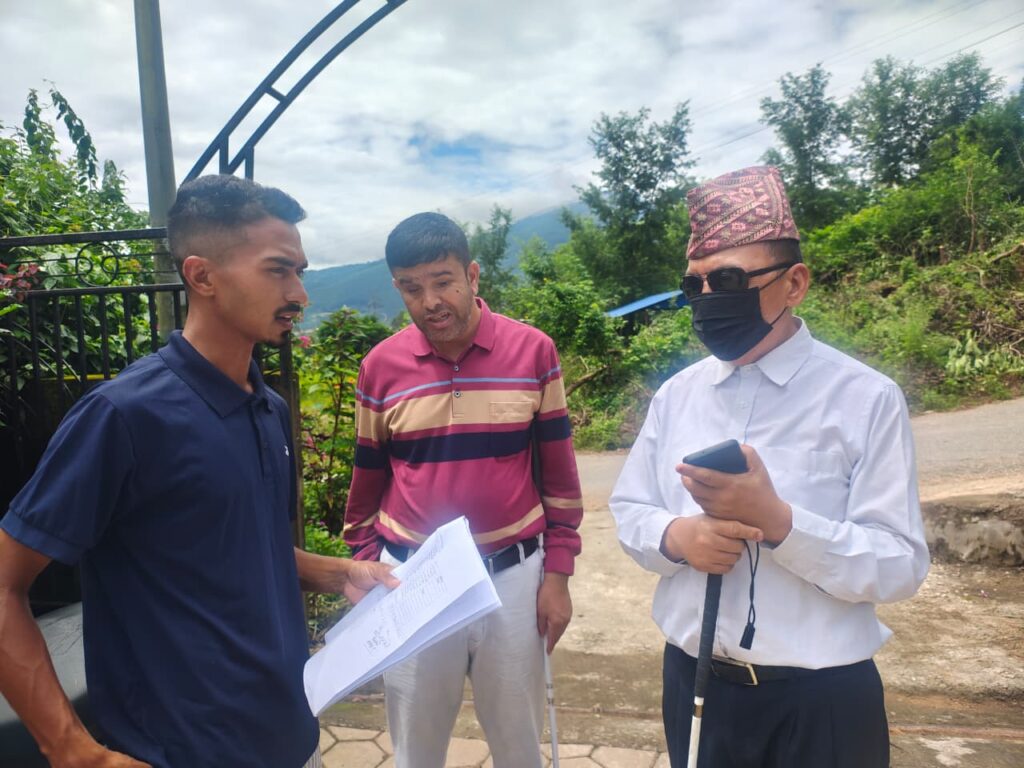 An outdoor setting showing three individuals (Representative of Church, Mr. Khom (Inclusion Advisor and Mr. Amrit Rai, Director of NAB) standing together near a gate, with one person holding papers while discussing them with the others.