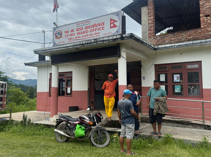 An outdoor setting showing the entrance of the 10 No. Ward Office building with a sign in Nepali and English, featuring several individuals gathered outside and a parked motorcycle in the foreground.