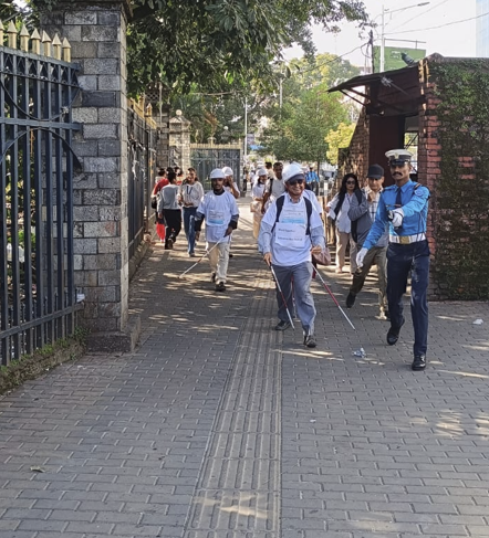 A group of participants wearing white t-shirts and white caps walk outdoors on a paved pathway using white cane mobility aids, guided by a traffic police officer in blue uniform.